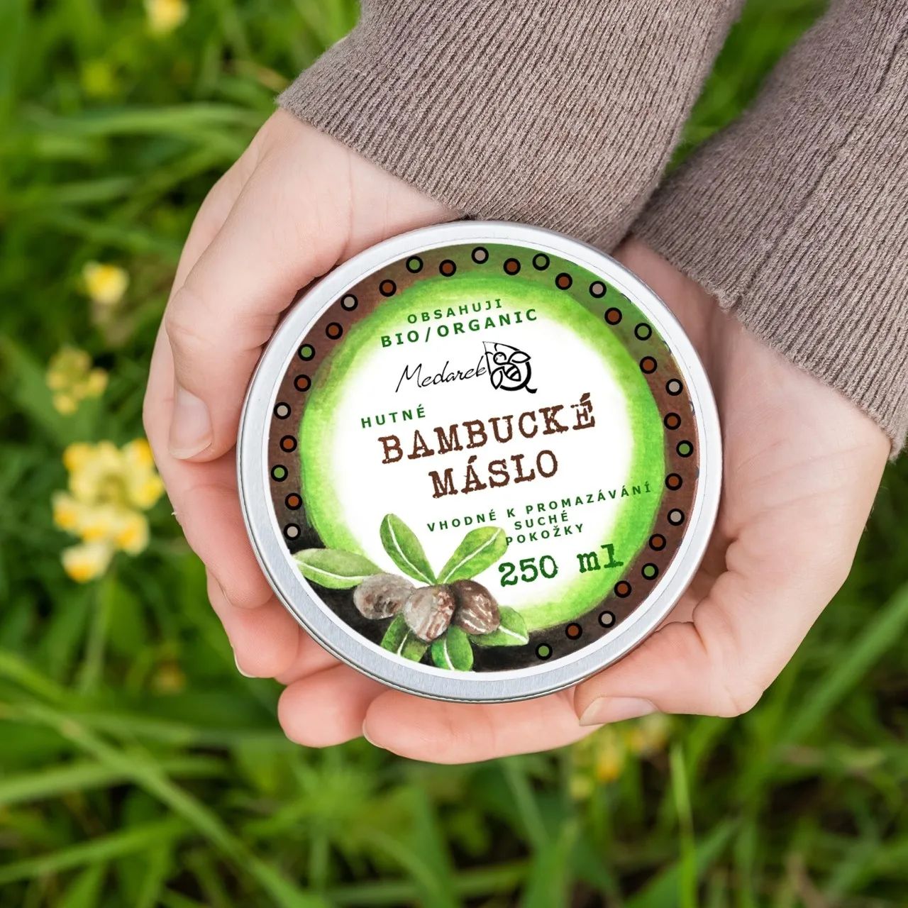 Hands holding a Medarek Bio Shea Butter (250 ml) tin over green grass.